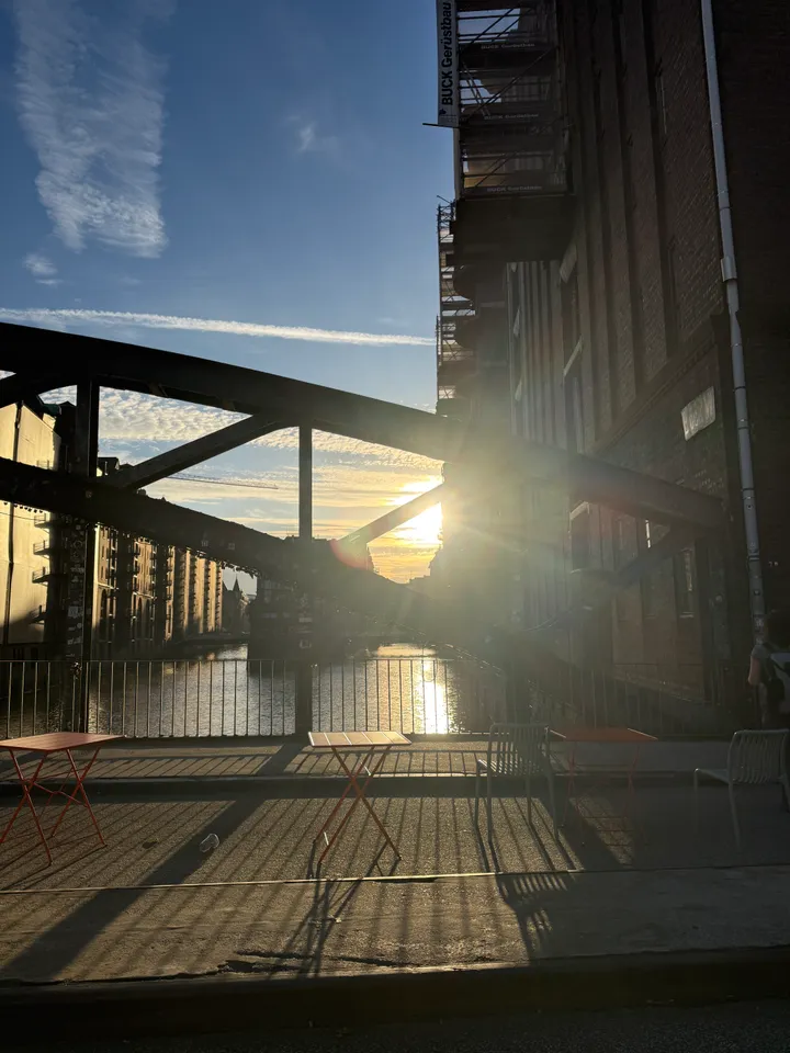 Rote Klapptische und Metallstühle auf einem Brückengehweg im Gegenlicht, dahinter Stahlbogen, Fleet und Speichergebäude