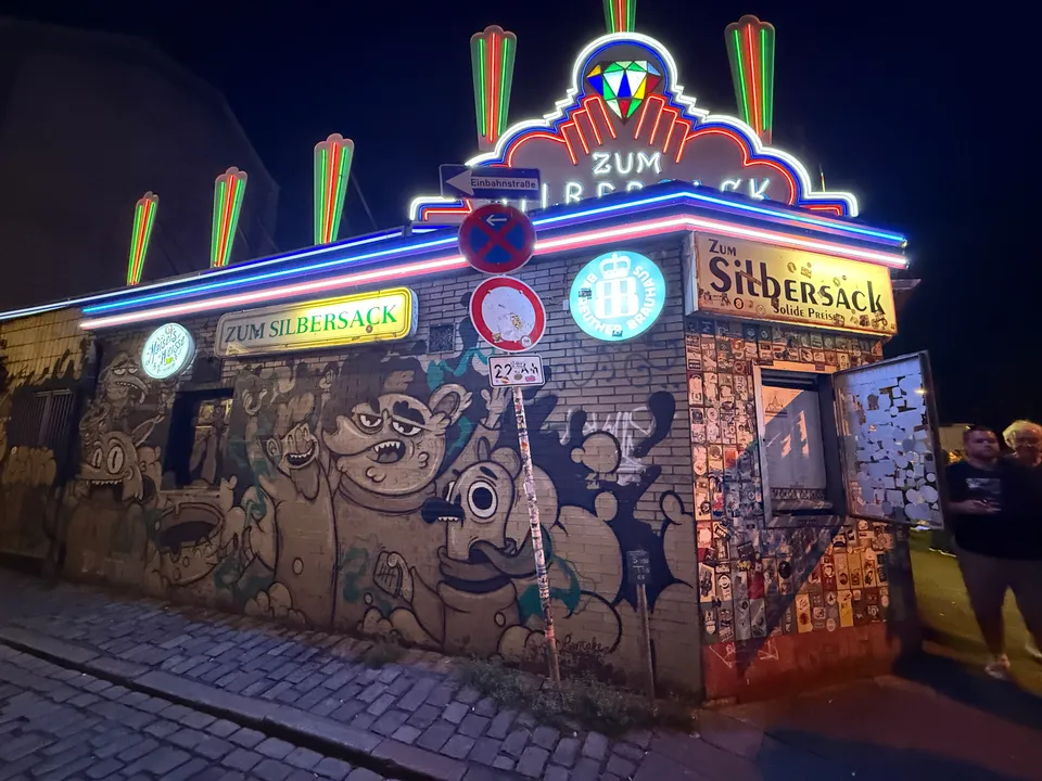 Backsteinfassade mit Comic-Wandbild, Leuchtschildern „Zum Silbersack“ und bunter Neon-Dachreklame bei Nacht