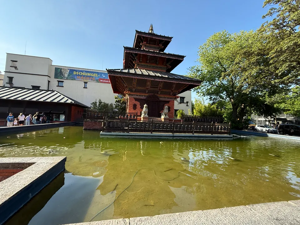 Nepalesische Pagode mit Löwenfiguren in einem Wasserbecken, dahinter ein helles Gebäude mit Werbebanner