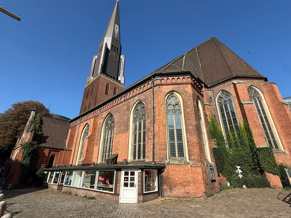 Rote Backsteinkirche mit hohem Turm, gotischen Spitzbogenfenstern, Efeu und flachem Ladenanbau an einem gepflasterten Platz