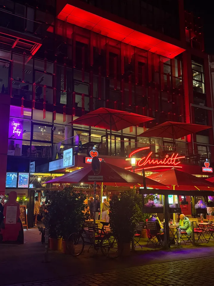 Rot beleuchtete Glasfassade mit Schmidt-Neonschriftzug, roten Sonnenschirmen und Außengastronomie bei Nacht