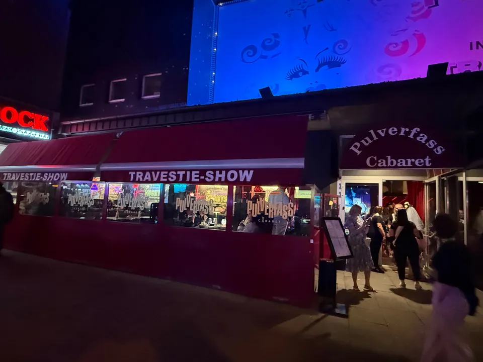 Roter Wintergarten mit Aufschrift Travestie-Show und Eingang Pulverfass Cabaret mit Gästen, nachts, blau angestrahltes Transparent