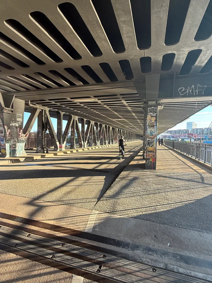 Fahrbahn und Fußweg unter einer Stahlbrücke mit Radfahrer, beklebtem Pfeiler und Graffiti-Stützen