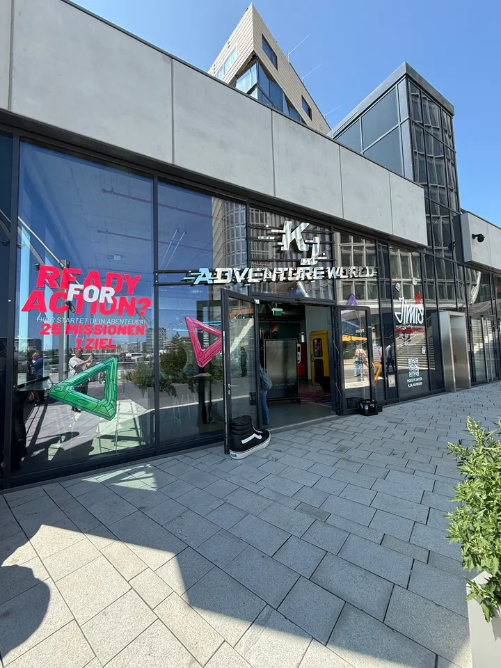 Glasfassade mit Schriftzug KJ Adventure World, offener Eingang, rotes Plakat „Ready for Action?“ und gepflasterter Vorplatz