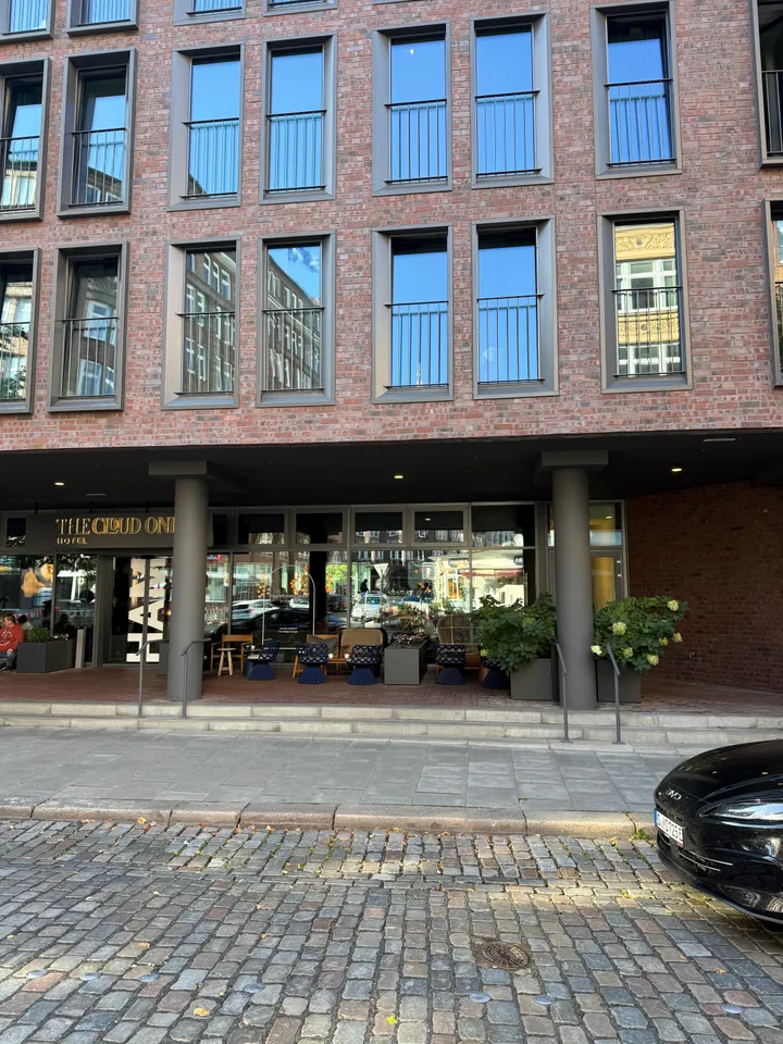Eingangsbereich eines Hotels aus Klinker mit Säulen, Außenterrasse und Pflanzkübeln an einer Kopfsteinpflasterstraße