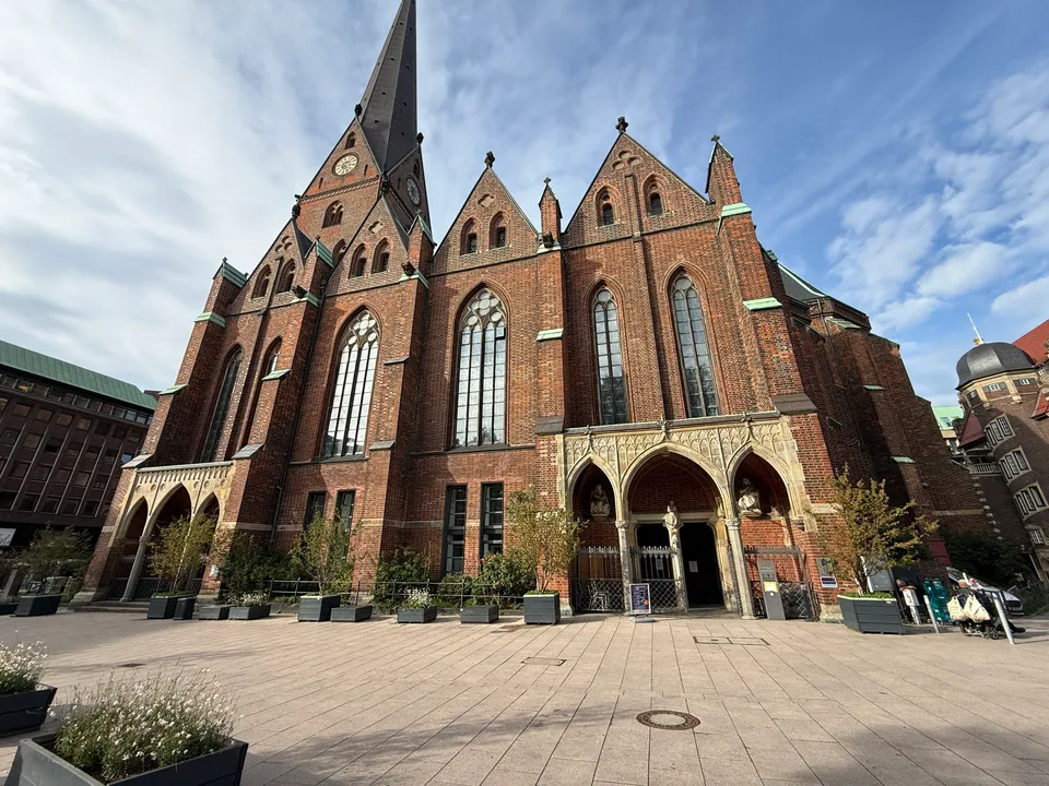 Giebelfassade einer neugotischen Backsteinkirche mit Sandsteinportal, Uhrturm und Pflanzkübeln auf gepflastertem Platz