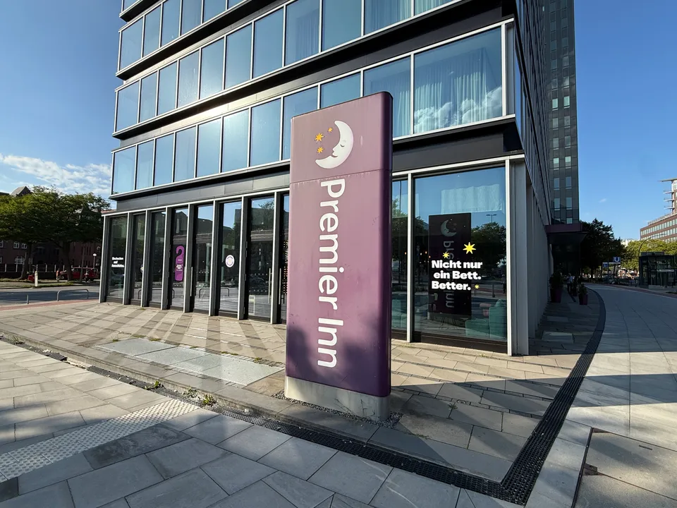 Violette Premier-Inn-Stele vor verglaster Hotelfassade mit Werbeslogan im Fenster und gepflastertem Gehweg bei Sonnenschein