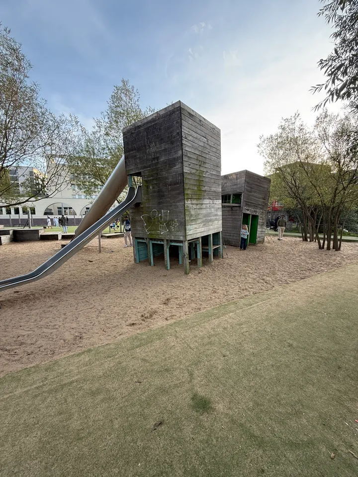 Hoher Holzspielturm auf Stelzen mit langer Metallrutsche und kleinem Holzhaus auf sandigem Spielplatz