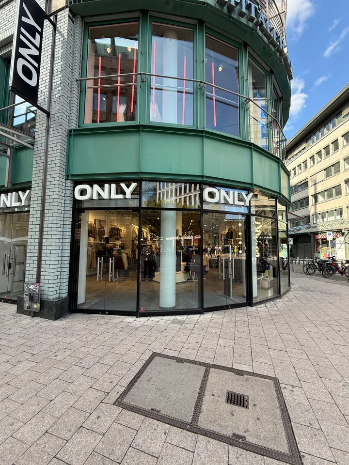 Runde Eckfassade eines ONLY-Modegeschäfts mit Glasfront, grünem Obergeschoss und gepflasterter Fußgängerzone davor