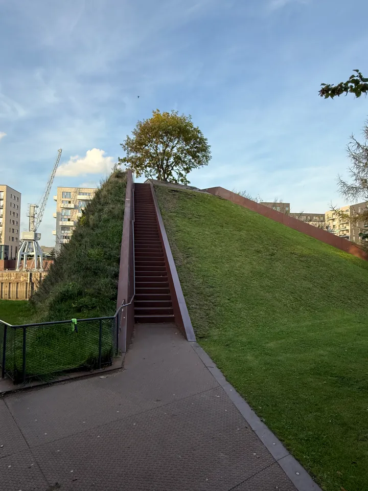 Steile Treppe mit rotbraunen Wangen führt einen Grashügel hinauf zu einem Baum, links ein Hafenkran