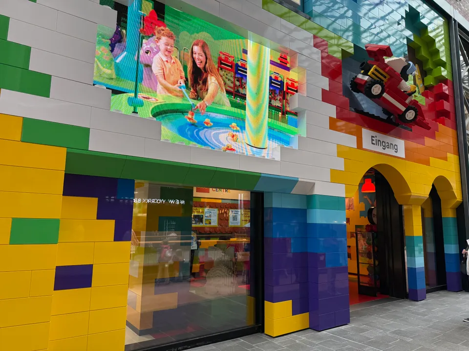 Bunte Fassade aus großen LEGO-Steinen mit Videowand, LEGO-Rennauto und Rundbogeneingang mit Schild „Eingang“