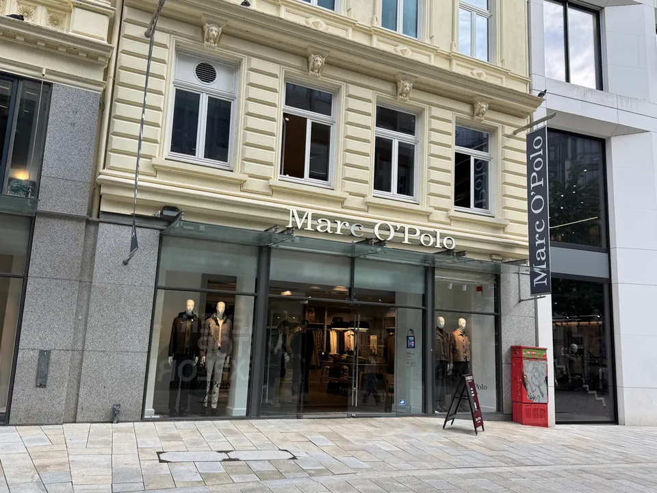 Marc-O'Polo-Geschäft im Erdgeschoss eines cremefarbenen Altbaus mit Glasvordach, Schaufensterpuppen und Kundenstopper in einer Fußgängerzone