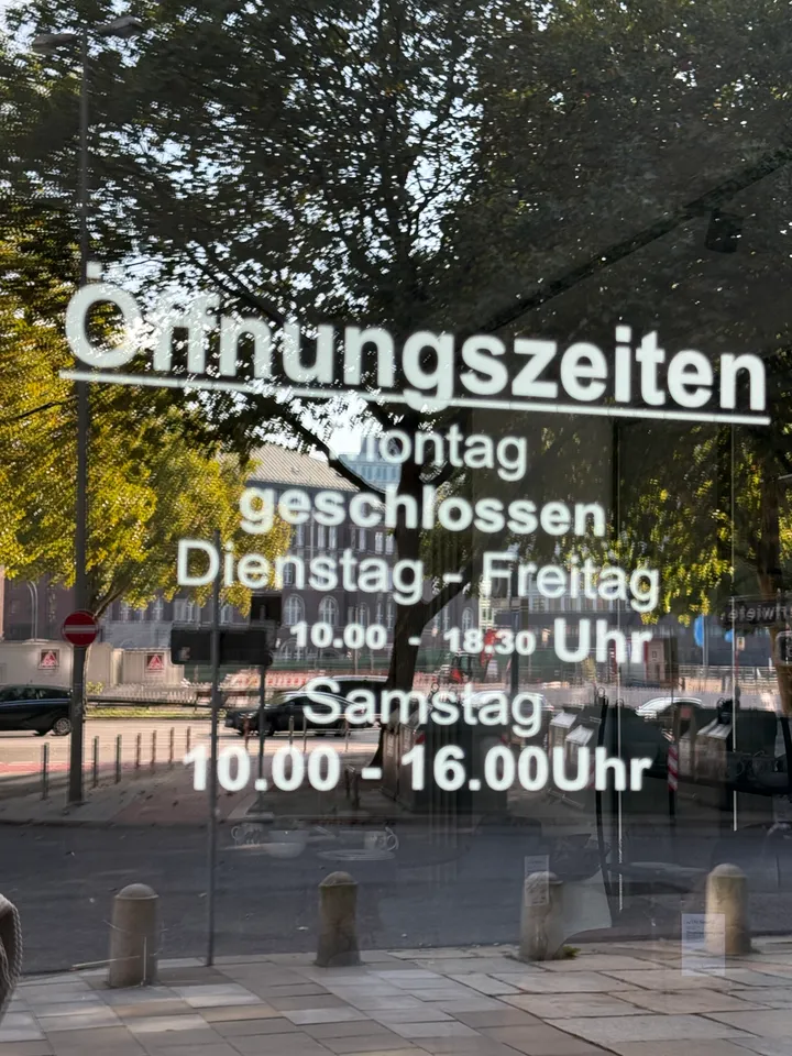 Schaufensterscheibe mit weißer Beschriftung der Öffnungszeiten, darin gespiegelte Bäume und Straße