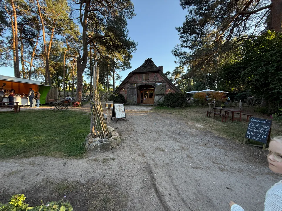 Giebel eines reetgedeckten Fachwerkhauses mit Torbogen, davor Sandplatz, Biertische und Tafel mit Öffnungszeiten
