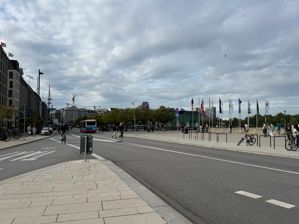 Breite Straße am Jungfernstieg mit Radfahrer und Bus, rechts Fahnenreihe an der Alsterpromenade, bewölkter Himmel