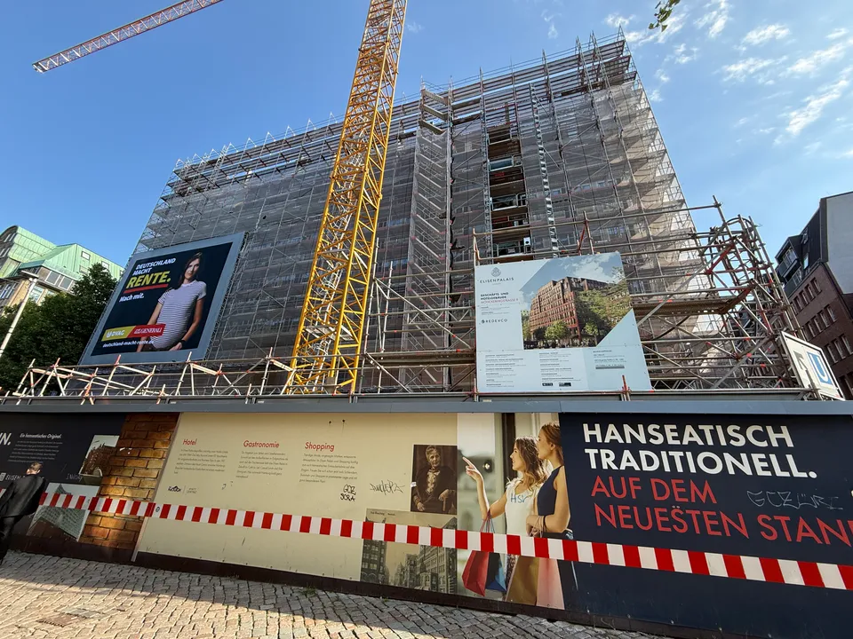 Eingerüsteter Neubau mit gelbem Kran, Bauschild, Werbeplakat und bedrucktem Bauzaun unter blauem Himmel