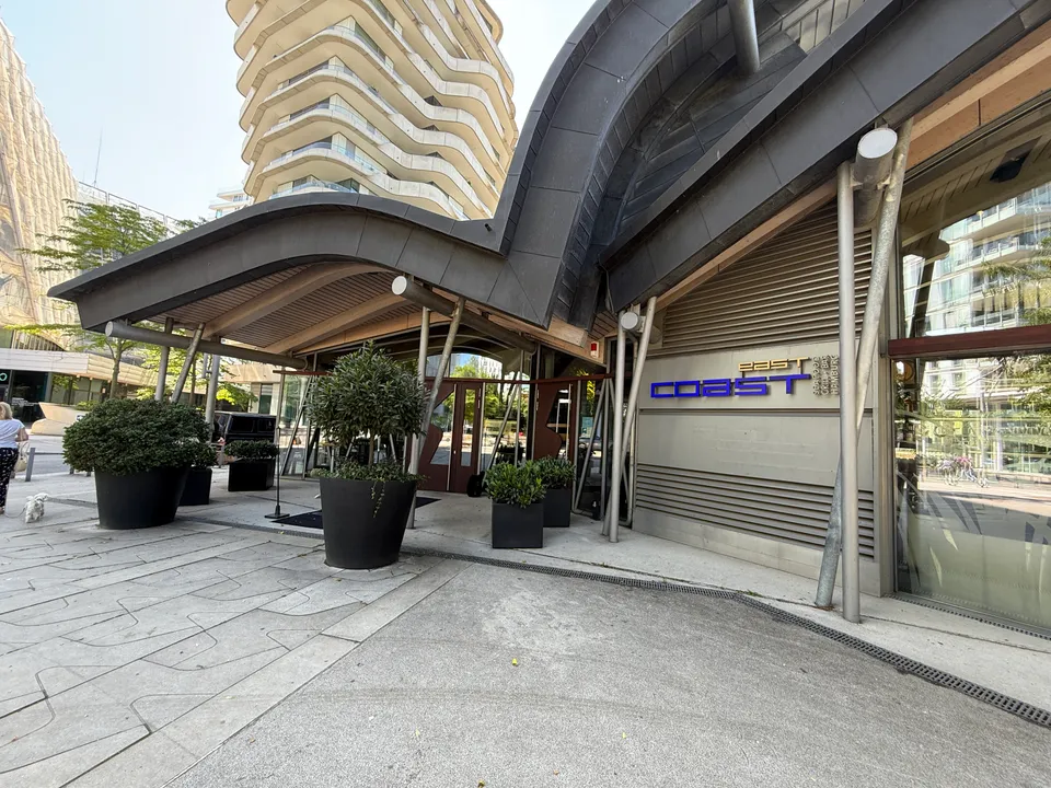 Eingang eines Restaurants unter geschwungenem Dach mit Schriftzug east coast, Pflanzkübel davor, dahinter ein Wohnhochhaus