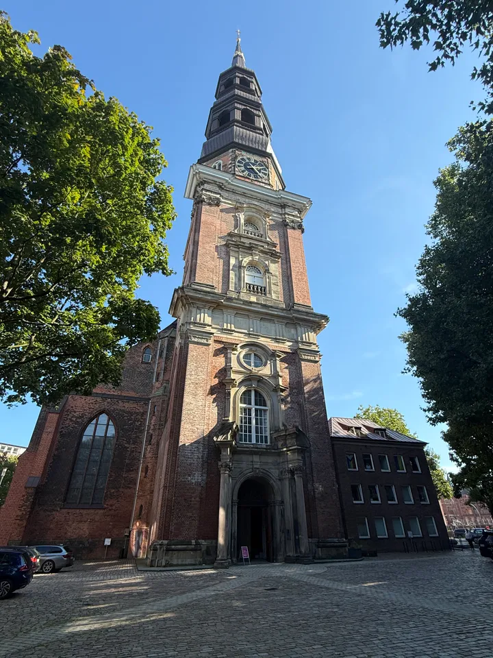 Barock geschmückter Backsteinkirchturm mit Rundbogenportal, Uhr und gestufter Turmhaube über einem gepflasterten Platz