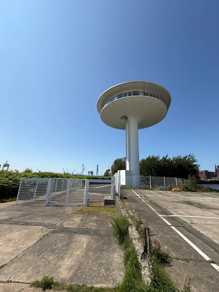 Weißer Turm mit scheibenförmigem Aufbau hinter Gitterzaun und Betonfläche, im Hintergrund Hafenkräne unter blauem Himmel