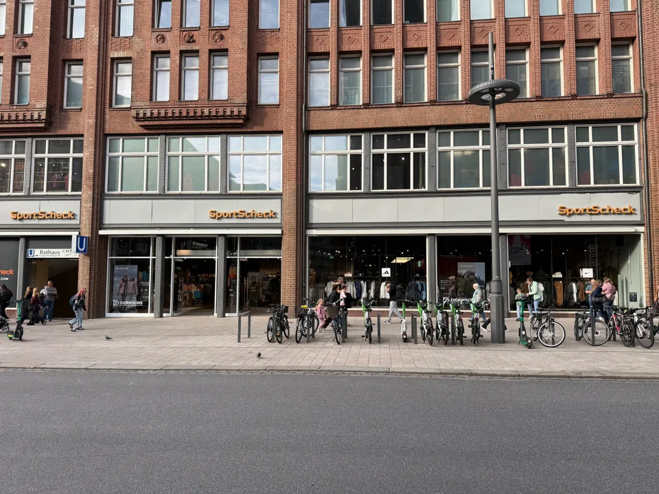 Backsteinfassade mit SportScheck-Schriftzug, Schaufenstern, U-Bahn-Eingang Rathaus und abgestellten Fahrrädern auf breitem Gehweg