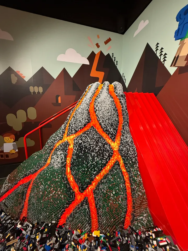 Vulkan aus LEGO-Steinen mit orangefarbenen Lavaströmen, roter Rutsche und gemalter Berglandschaft an der Wand