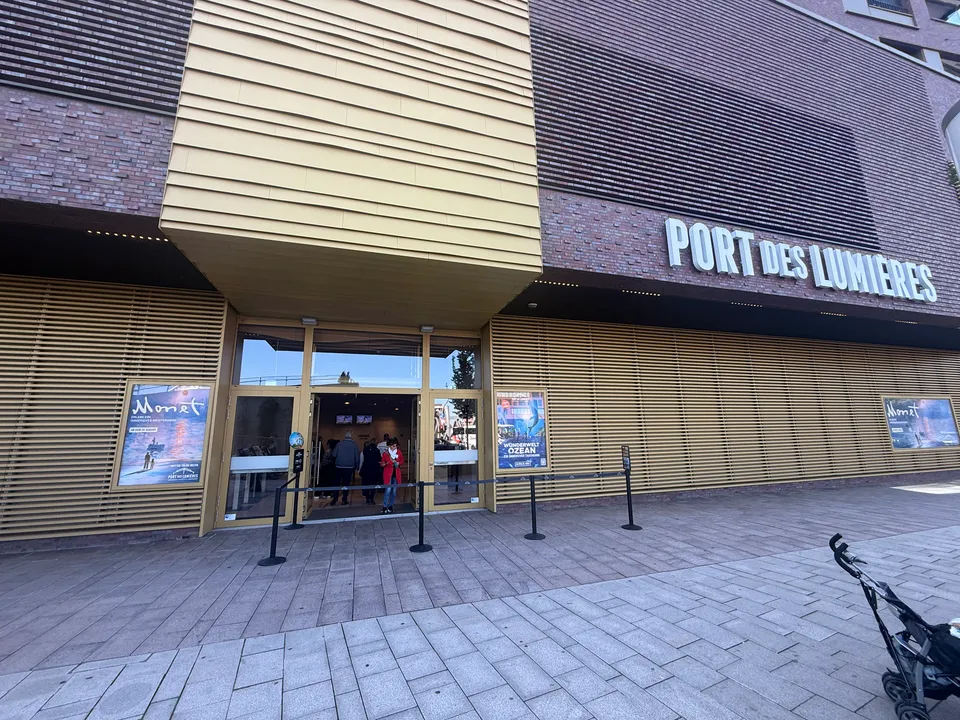 Eingang des Port des Lumières mit goldener Lamellenfassade, Klinkerwand, Ausstellungsplakaten und Absperrbändern auf dem gepflasterten Vorplatz