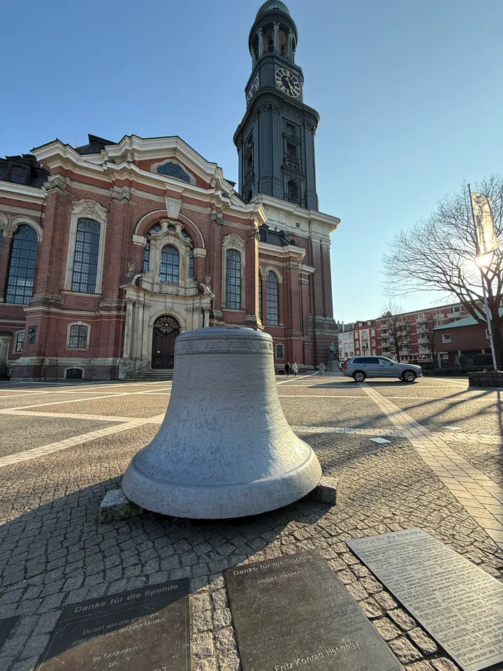 Glockenskulptur aus Stein mit Bodenplatten vor barocker Backsteinkirche mit hohem Uhrturm
