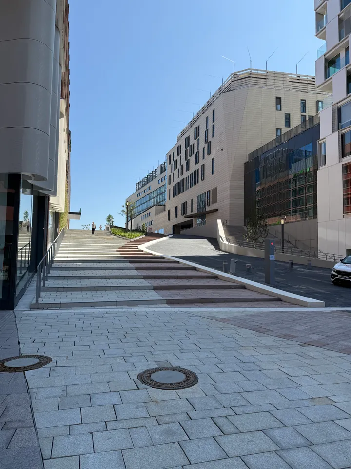 Breite Steintreppe und ansteigende Rampe zwischen modernen Gebäuden mit hellbeiger Fassade unter blauem Himmel