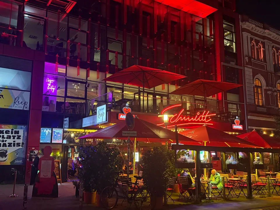 Rot beleuchtete Theaterfassade mit Schmidt-Neonschrift, Sonnenschirmen, Außentischen und angrenzendem Altbau bei Nacht