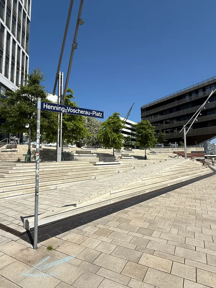 Straßenschild Henning-Voscherau-Platz vor breiter heller Treppenanlage mit Bäumen, schrägen Lichtmasten und dunklem Bürogebäude