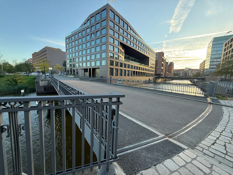 Gebogene Straßenbrücke über einen Kanal, dahinter ein Klinker-Bürogebäude und tiefstehende Sonne über dem Wasser