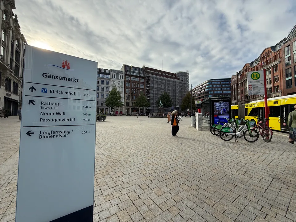 Wegweiserstele am Gänsemarkt mit Richtungsangaben, dahinter gepflasterter Platz, Bushaltestelle, Leihfahrräder und Geschäftshäuser
