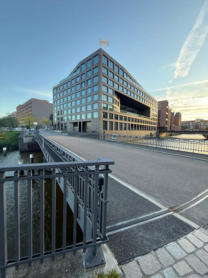 Straßenbrücke mit grauem Metallgeländer über einen Kanal, dahinter ein großes Bürogebäude aus dunklem Klinker