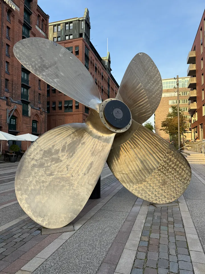 Große messingfarbene Schiffsschraube auf gepflastertem Platz vor Backsteinspeichern und modernen Wohnhäusern in der HafenCity