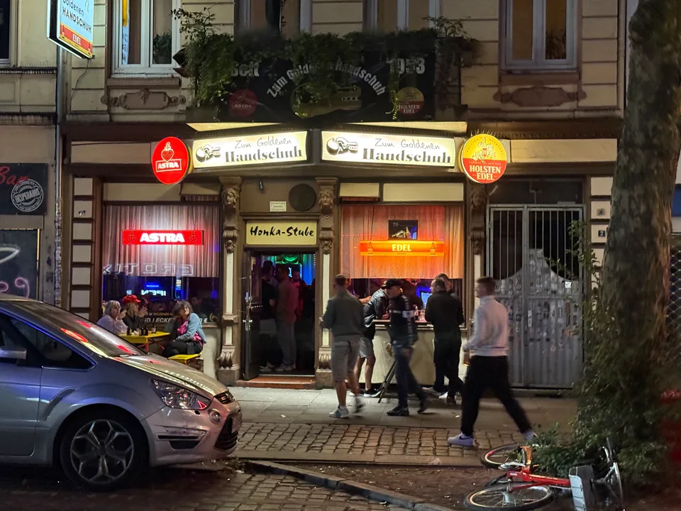 Beleuchtete Fassade der Kneipe Zum Goldenen Handschuh bei Nacht, mit Gästen vor dem Eingang und parkendem Auto