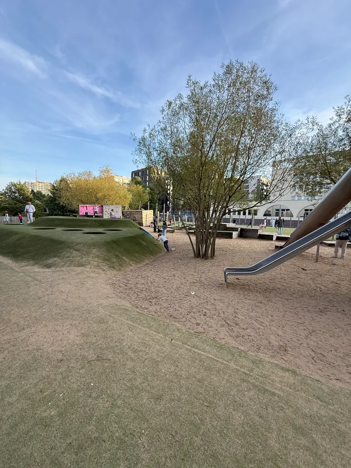 Kunstrasenhügel mit runden Öffnungen neben Sandfläche, Baum und Metallrutsche auf großem Stadtspielplatz unter blauem Himmel