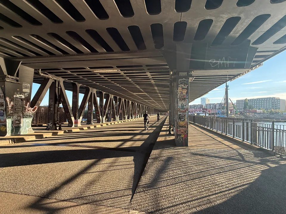 Fahrbahn unter einer Stahlbrücke mit V-Stützen, Radfahrenden, Fußweg am Geländer und Wasser mit Neubauten rechts