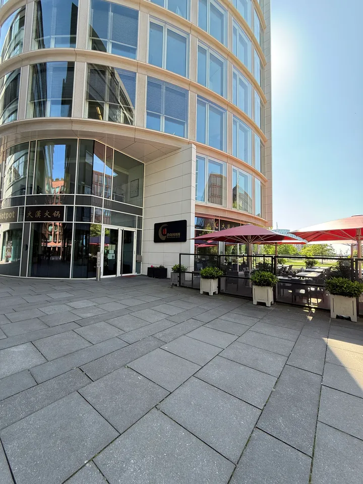 Abgerundetes Bürogebäude mit Eingang des Dahan Restaurant und Hotpot, rechts Terrasse mit roten Sonnenschirmen