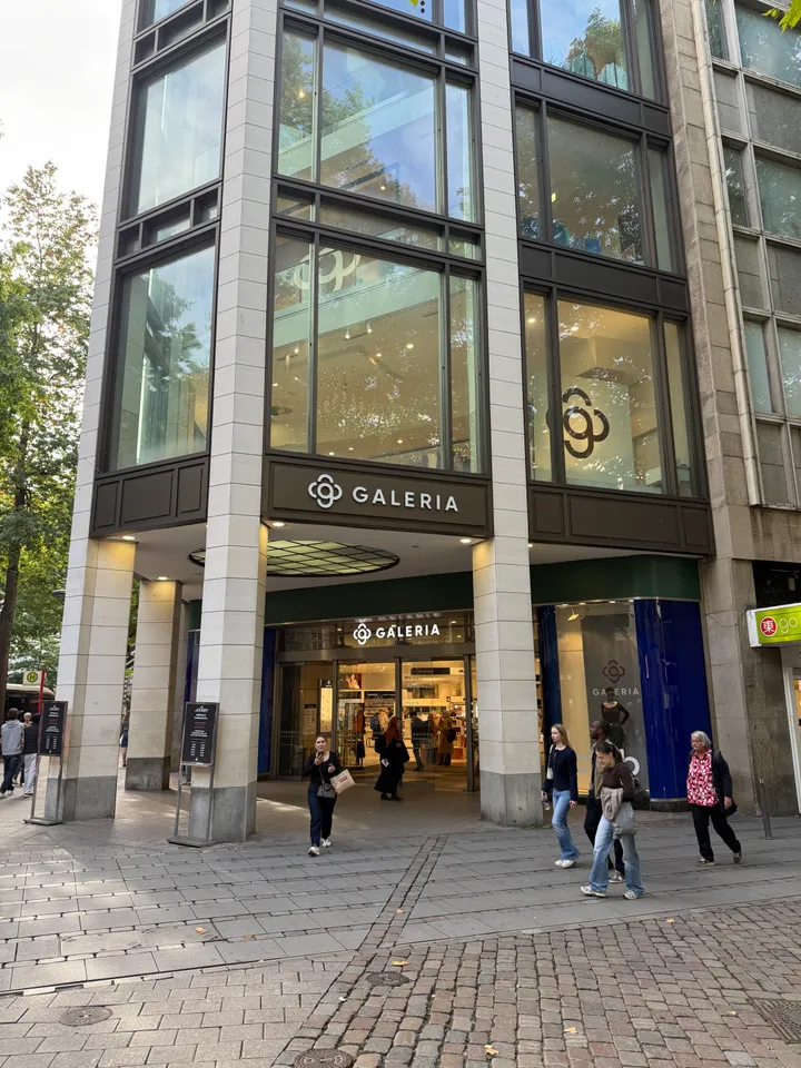 Eckeingang des GALERIA-Kaufhauses mit Steinpfeilern und großen Fenstern, davor Passanten auf gepflastertem Gehweg
