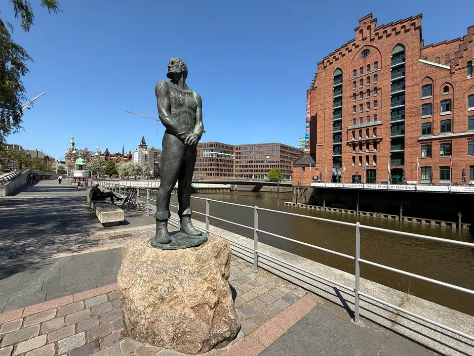 Bronzestatue Klaus Störtebekers mit gefesselten Händen auf Granitsockel, dahinter Hafenbecken und Maritimes Museum Hamburg