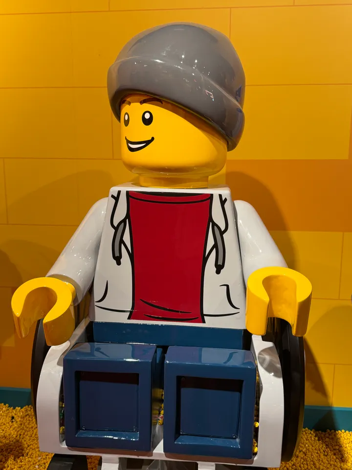 Große LEGO-Minifigur mit grauer Mütze und weißer Jacke sitzt im Rollstuhl vor gelber Wand