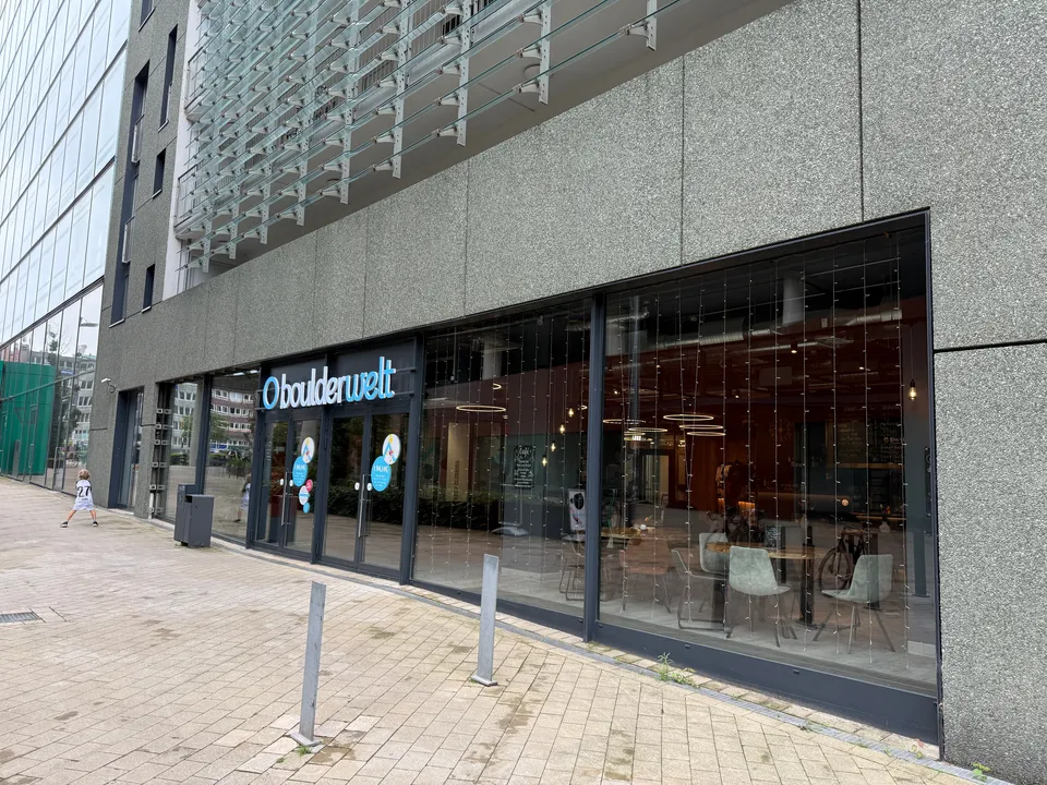 Glasfront im Erdgeschoss eines grauen Gebäudes mit Schriftzug boulderwelt über dem Eingang, davor ein gepflasterter Gehweg mit Pollern