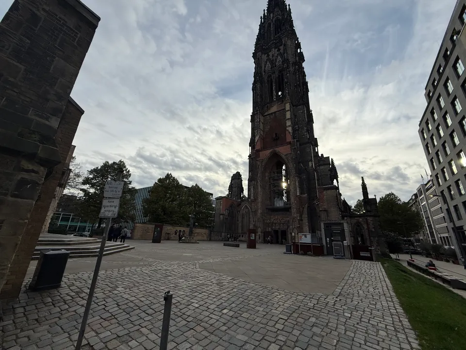 Gepflasterter Platz vor dem hohen Turm der Kirchenruine St. Nikolai, links Mauerpfeiler und Hinweisschilder