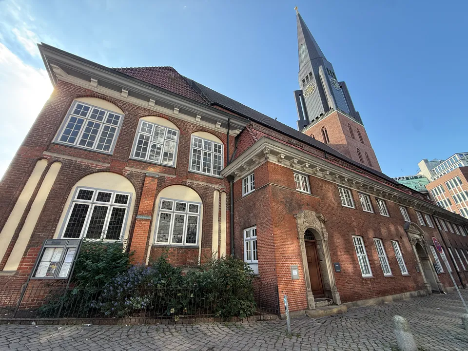 Backsteinbau mit großen Rundbogenfenstern und Sandsteinportal, dahinter ein Kirchturm mit Uhr und spitzem Helm