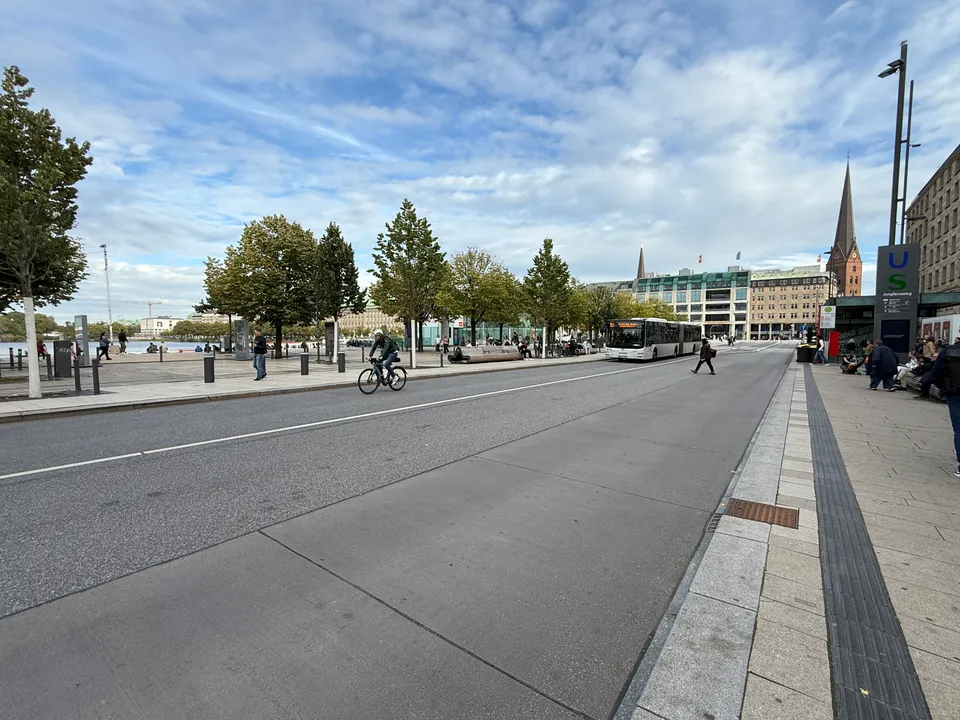 Jungfernstieg mit Radfahrer und Gelenkbus, links Bäume an der Binnenalster, rechts U- und S-Bahn-Schild, Kirchturm