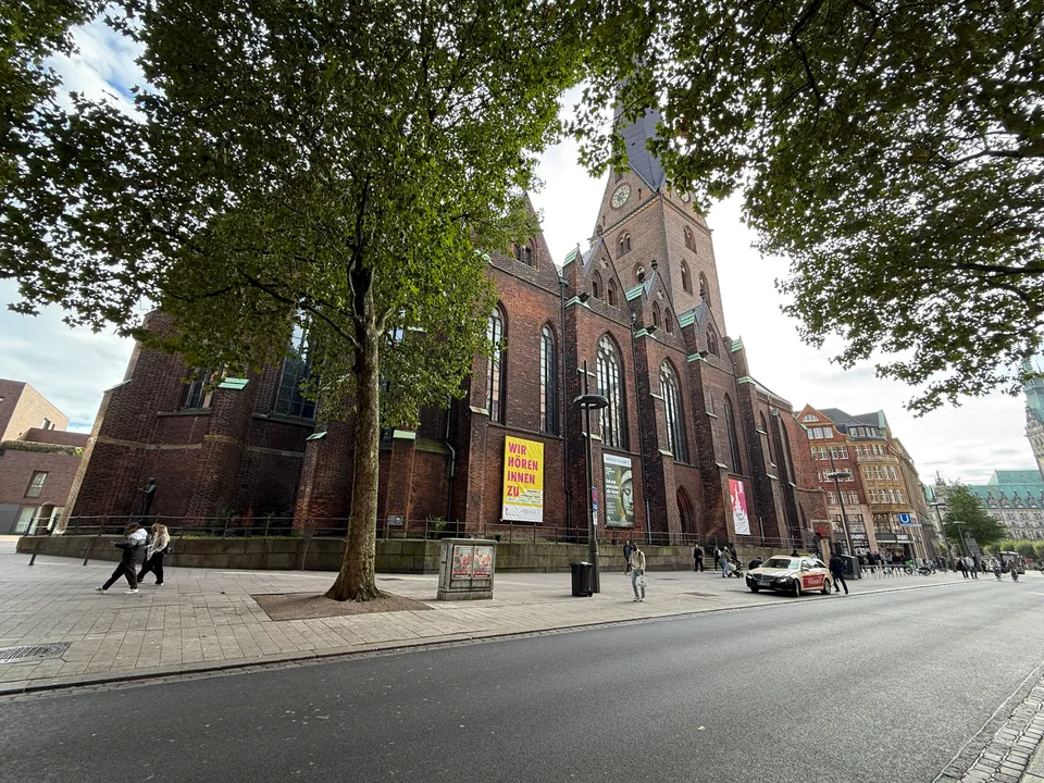 Backsteinkirche mit hohem Turm und gelbem Banner hinter Bäumen an einer Straße, rechts das Rathaus