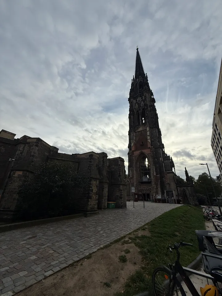 Neugotischer Kirchturm der Ruine St. Nikolai hinter Mauerresten, davor Pflasterweg, Rasen und Fahrrad unter bewölktem Himmel