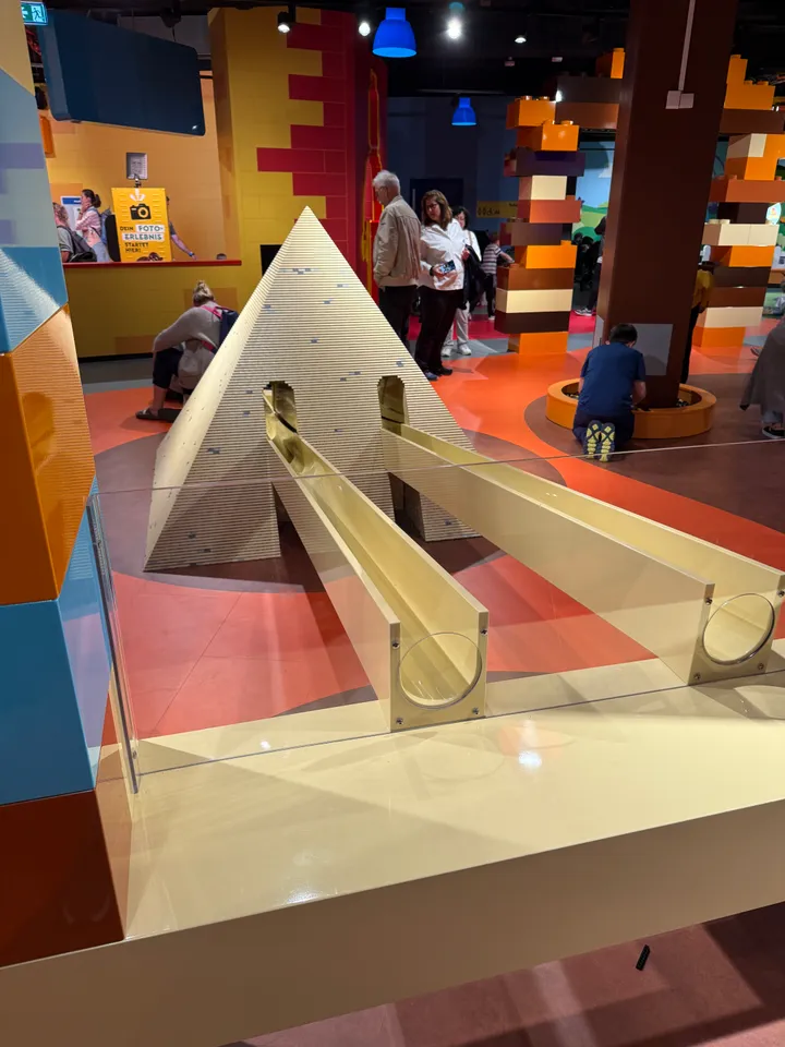 Beigefarbene LEGO-Pyramide mit zwei Rollbahnen in einer Vitrine, dahinter eine Spielhalle mit Besuchern