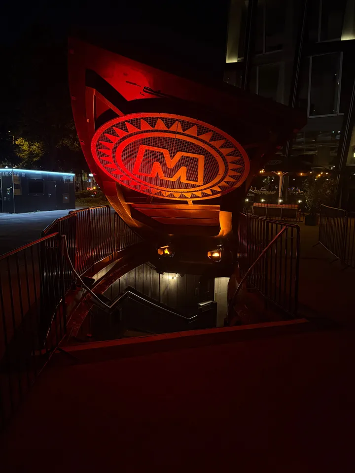 Rot-orange beleuchteter, aufgeklappter Clubeingang mit ovalem M-Logo und nach unten führender Treppe bei Nacht