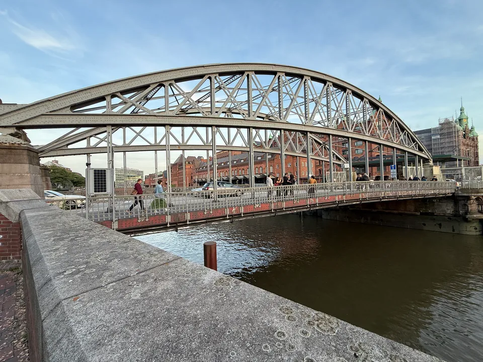 Stählerne Bogenbrücke über einen Kanal, dahinter rote Backsteinspeicher und ein Gebäude mit grünen Türmen
